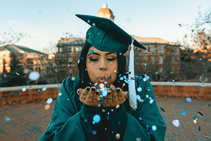 Woman blowing confetti wearing a green graduation cap and gown with a college in the background.