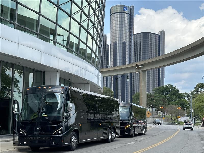 Two black luxury motor coaches parked outside a modern building in downtown Detroit, representing basketball transportation for the Detroit Pistons.